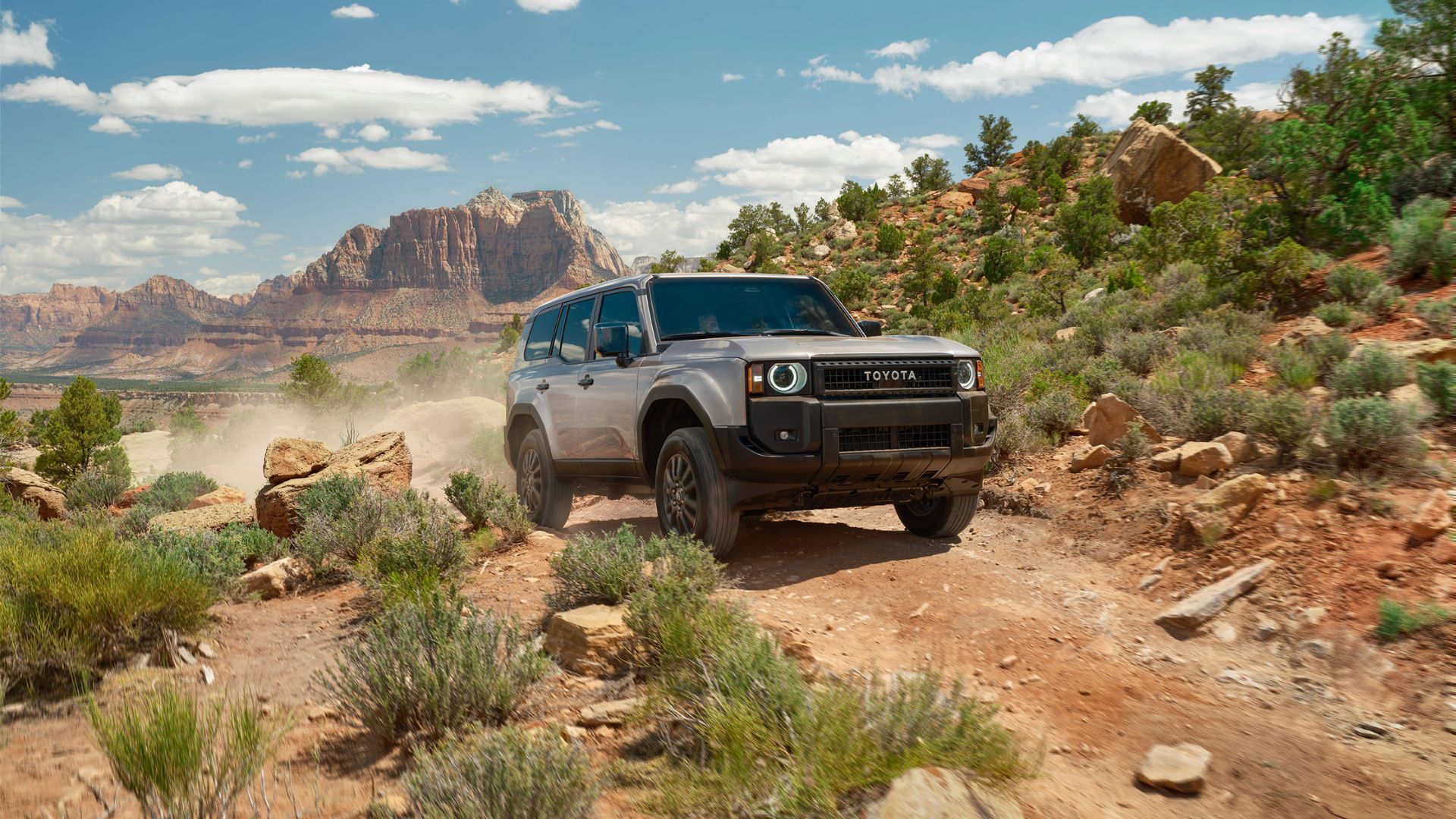 Friont 3/4 view of a silver 2025 Toyota Land Cruiser off-roading in the desert with mountains in the background.