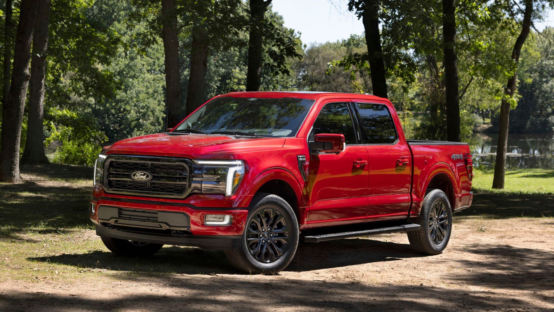 Static front 3/4 shot of a red 2024 Ford F-150 Lariat parked on dirt with trees in the background.