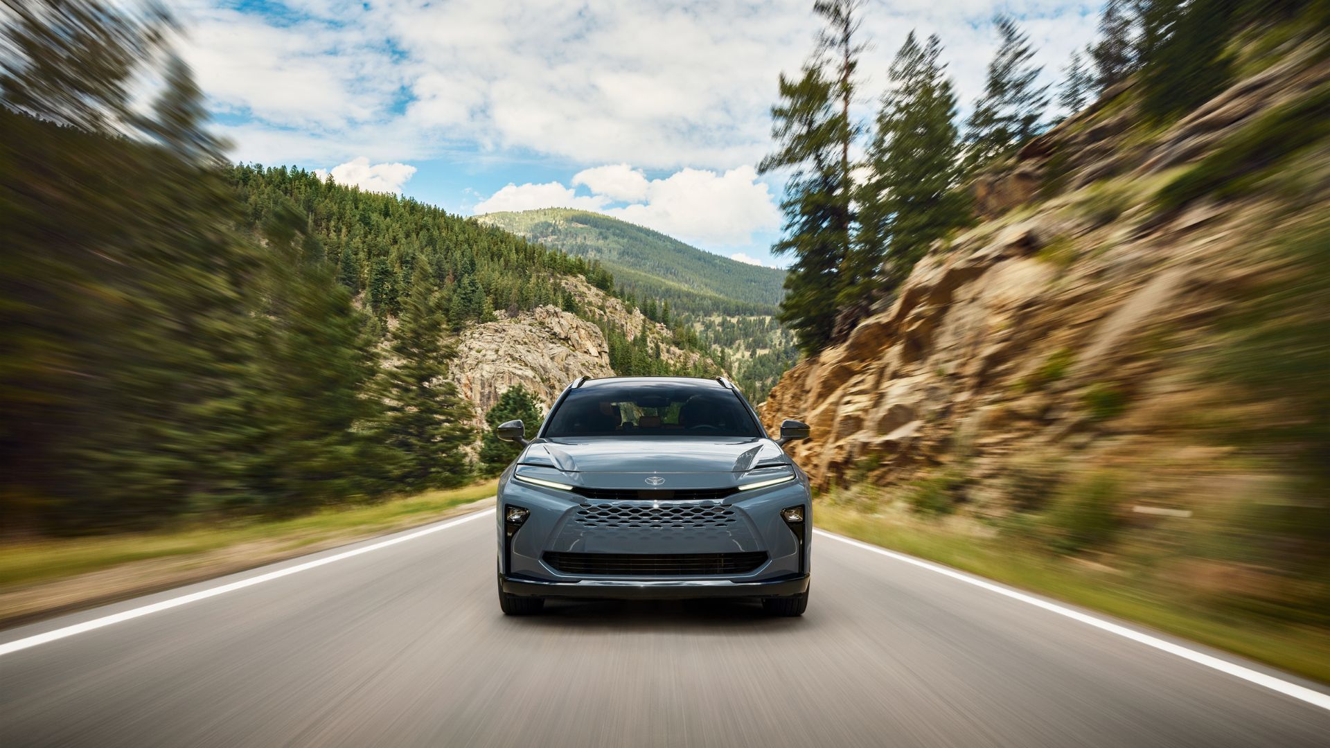 Dynamic front-end shot of a gray 2025 Toyota Crown Signia driving on a country road lined by trees a mountain in the background.