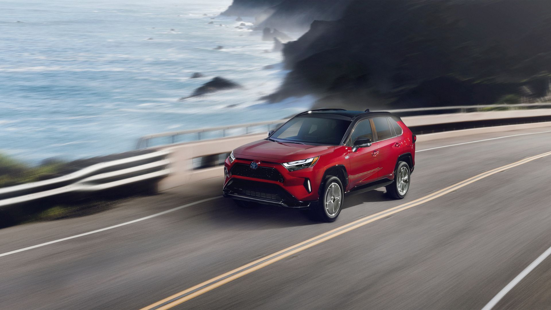 Dynamic front 3/4 shot of a red 2024 Toyota RAV4 Prime XSE driving on a road running beside the ocean.