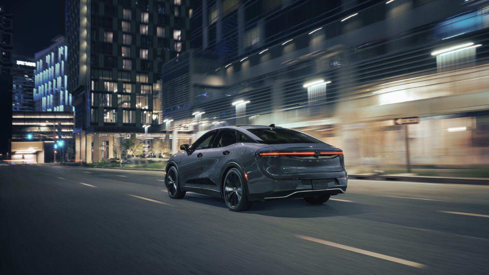 Dynamic rear 3/4 shot of a gray 2026 Toyota Crown Nightshade driving at night in a city.