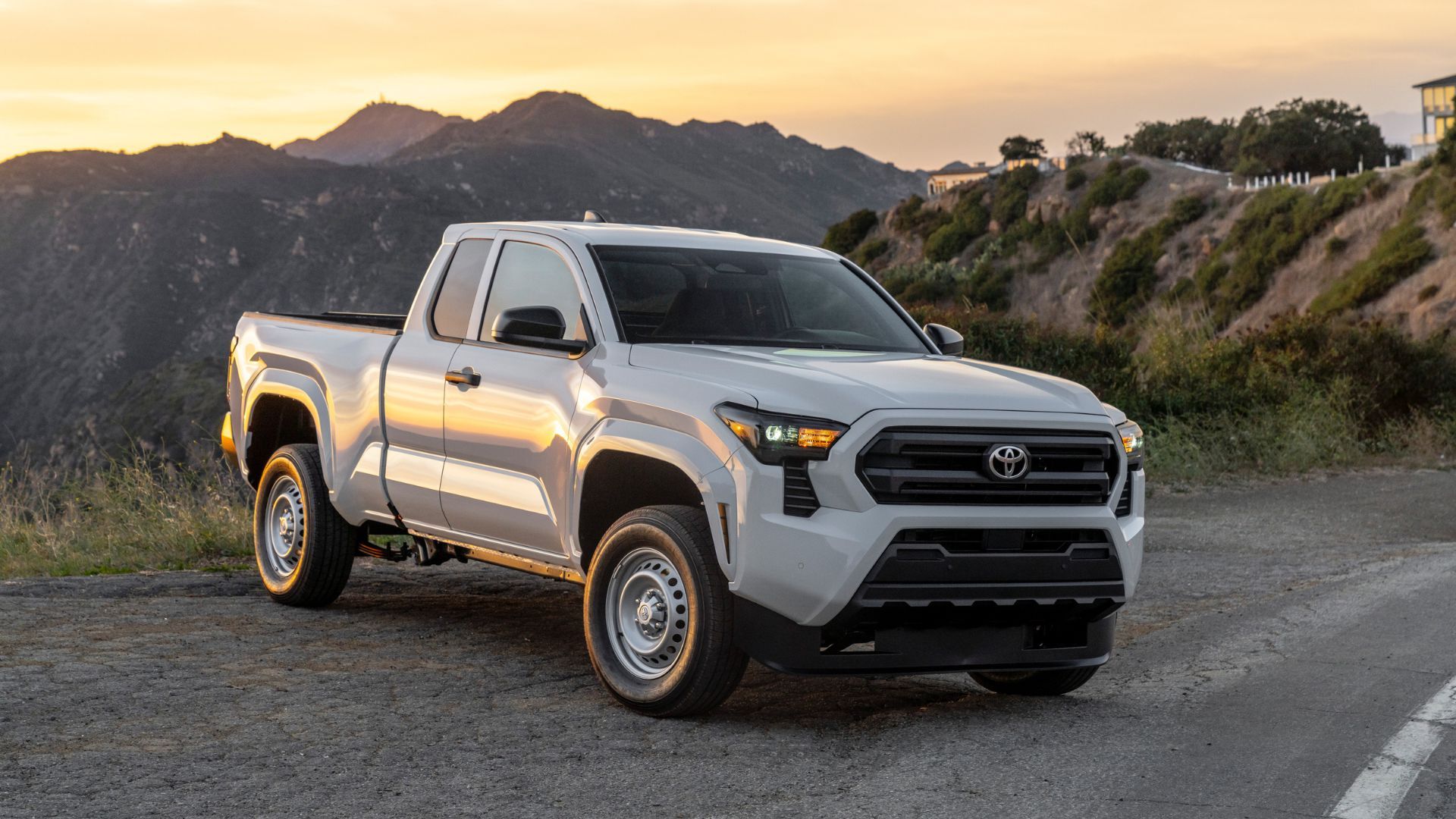 Static front 3/4 shot of a white 2025 Toyota Tacoma SR with mountains and a property in the background.