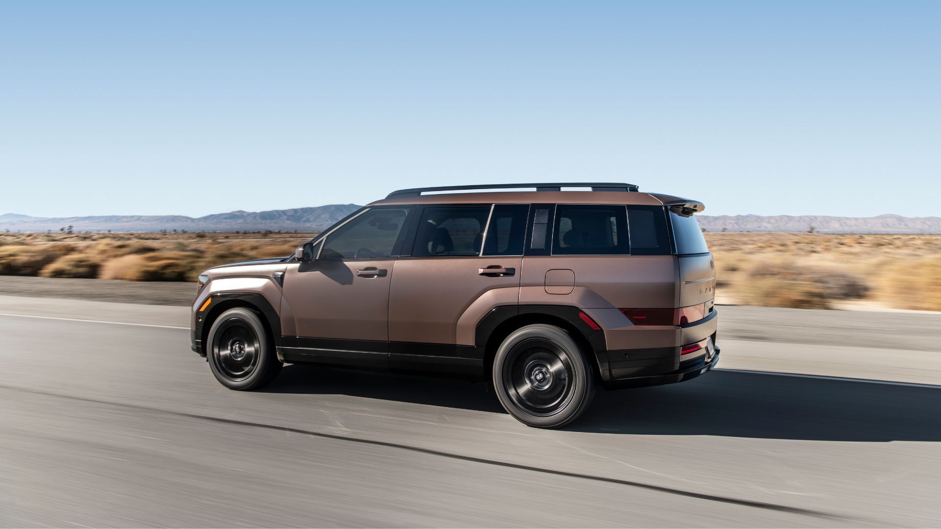 Dynamic side profile shot of a bronze 2025 Hyundai Santa Fe Hybrid driving on a country road through the desert.