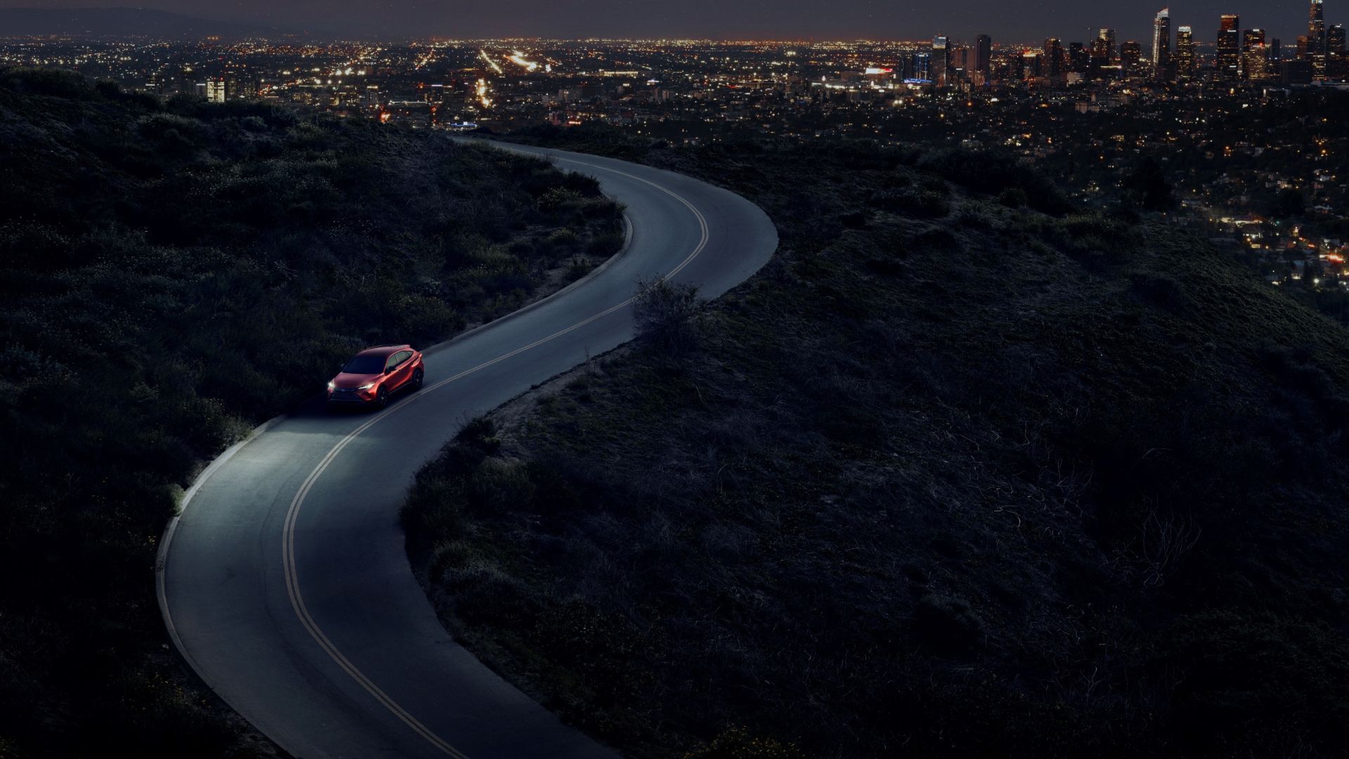 Dynamic overhead front 3/4 shot of a red 2024 Toyota Venza Nightshade driving on a winding road.