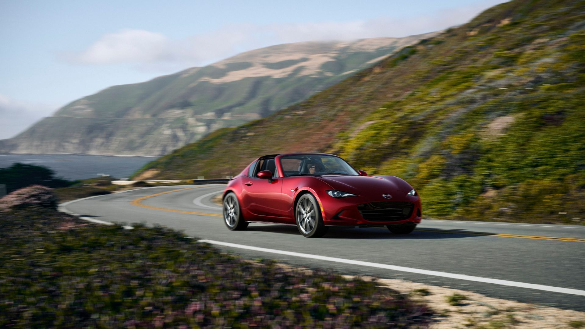Front 3/4 shot of a red 2024 Mazda MX-5 Miata RF driving on a winding road with the ocean in the background.